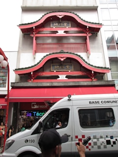 A traditional Asian-style building with red and green decorative elements is set against a modern urban backdrop. Two ornate, curved eaves are prominent, and there are signs with Asian characters. Below, a white vehicle labeled 'BASE COMUNITÁRIA' is passing by, with a man in a cap holding an object that emits bubbles. Several people can be seen near the entrance of a shop.