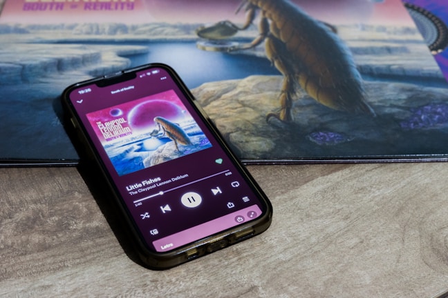 A smartphone is displaying a music application with a song named 'Little Fishes' by The Claypool Lennon Delirium. The phone screen shows album art featuring a cosmic scene with a large creature overlooking a water body. The background contains a similar artwork involving a surrealistic landscape with a creature.