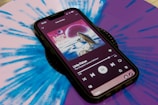 A smartphone displays a music player interface showing album art and song details on its screen. The phone is placed on a vibrant blue and purple vinyl record, which has a kaleidoscope-like pattern. The album cover features a psychedelic landscape with a celestial theme.