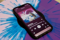 A smartphone displays a music player interface showing album art and song details on its screen. The phone is placed on a vibrant blue and purple vinyl record, which has a kaleidoscope-like pattern. The album cover features a psychedelic landscape with a celestial theme.