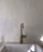 A brass kitchen faucet stands against a textured, off-white plaster wall. The faucet rises from a white ceramic sink and casts a subtle shadow. Natural light creates soft highlights on the wall and faucet, enhancing the rustic texture of the plaster.