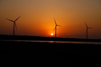 A serene landscape featuring three wind turbines silhouetted against a vibrant sunset. The sun is setting near the horizon, casting an orange glow across the sky and reflecting in a body of water beneath the turbines.