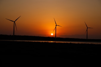A serene landscape featuring three wind turbines silhouetted against a vibrant sunset. The sun is setting near the horizon, casting an orange glow across the sky and reflecting in a body of water beneath the turbines.
