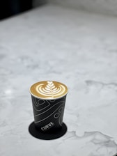 A cup of latte with intricate latte art sits on a marble surface. The cup is black with a unique white pattern and the word 'CURVE' printed on it.
