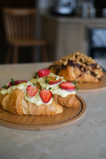 Two pastries are placed on wooden trays. The foreground pastry is a croissant topped with cream, sliced strawberries, and a sprinkling of green garnish. The background pastry appears to be a bread topped with nuts and chocolate drizzle. The setting includes a blurred background with a chair and counter visible.