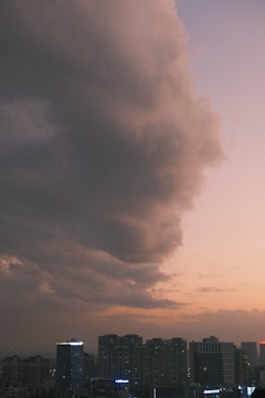 A skyline view featuring tall buildings with lights on, under a vast, dramatic sky filled with large, dark clouds. The horizon displays a soft gradient from pink to purple, suggesting either a sunrise or sunset.