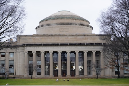 A neoclassical building features a prominent dome and a row of columns at the entrance. The facade includes large windows and is surrounded by leafless trees, suggesting a winter setting. The stone structure is adorned with architectural details typical of educational institutions.
