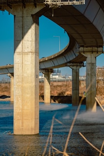 An elevated highway structure with large concrete pillars supports the road above a body of water. The scene includes a clear sky and visible urban elements in the background. The sunlight casts shadows on the pillars, and there is some vegetation along the water's edge.