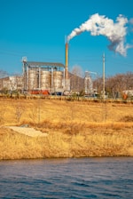 An industrial facility with large metal structures and a tall smokestack emitting white smoke into the clear blue sky. In the foreground, a grassy field with dry, golden-brown vegetation and a body of water create a natural contrast to the industrial scene.