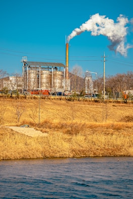 An industrial facility with large metal structures and a tall smokestack emitting white smoke into the clear blue sky. In the foreground, a grassy field with dry, golden-brown vegetation and a body of water create a natural contrast to the industrial scene.