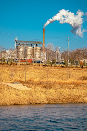 An industrial facility with large metal structures and a tall smokestack emitting white smoke into the clear blue sky. In the foreground, a grassy field with dry, golden-brown vegetation and a body of water create a natural contrast to the industrial scene.
