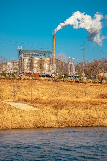 An industrial facility with large metal structures and a tall smokestack emitting white smoke into the clear blue sky. In the foreground, a grassy field with dry, golden-brown vegetation and a body of water create a natural contrast to the industrial scene.