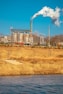 An industrial facility with large metal structures and a tall smokestack emitting white smoke into the clear blue sky. In the foreground, a grassy field with dry, golden-brown vegetation and a body of water create a natural contrast to the industrial scene.