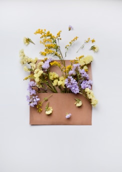 An open brown envelope is placed against a plain white background, filled with a variety of colorful flowers. Yellow, purple, and light cream blooms emerge from the envelope, with the flowers and some petals artistically scattered around.
