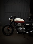 A Royal Enfield motorcycle is parked in front of a large, industrial-style metal door. The bike features a white and red fuel tank with chrome engine components and black tires. The background is dimly lit, highlighting the motorcycle's classic design.