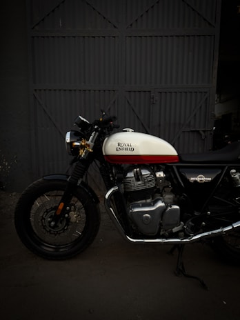 A Royal Enfield motorcycle is parked in front of a large, industrial-style metal door. The bike features a white and red fuel tank with chrome engine components and black tires. The background is dimly lit, highlighting the motorcycle's classic design.