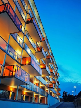 A modern apartment building with multiple glass balconies illuminated warmly at dusk. The perspective emphasizes the vertical lines, contrasted against a clear blue sky. Streetlamps line the adjacent road, and the horizon shows silhouettes of trees.
