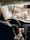 A person is driving a car viewed from the back seat, with a close-up of their hand on the steering wheel. The windshield appears dusty or wet, reflecting sunlight and partially obscuring the view of the road and other vehicles.