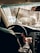 A person is driving a car viewed from the back seat, with a close-up of their hand on the steering wheel. The windshield appears dusty or wet, reflecting sunlight and partially obscuring the view of the road and other vehicles.
