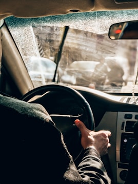 A person is driving a car viewed from the back seat, with a close-up of their hand on the steering wheel. The windshield appears dusty or wet, reflecting sunlight and partially obscuring the view of the road and other vehicles.