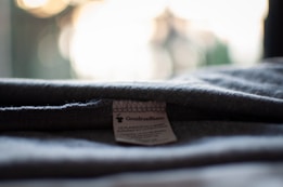 A close-up view of a garment label sewn onto grey fabric. The label has the text 'GoudronBlanc' and some additional information in small print. The fabric appears folded, and the background is blurred with warm and light tones.