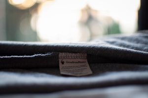 A close-up view of a garment label sewn onto grey fabric. The label has the text 'GoudronBlanc' and some additional information in small print. The fabric appears folded, and the background is blurred with warm and light tones.