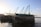 A large wooden boat is docked by the shore, with several masts and ropes extending upwards. The name 'MPIRAPESA' is visible on the side of the boat. The surrounding setting is calm, with the water reflecting the structures and the sky showing subtle tones of a sunset.