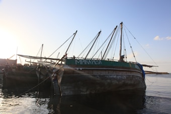 A large wooden boat is docked by the shore, with several masts and ropes extending upwards. The name 'MPIRAPESA' is visible on the side of the boat. The surrounding setting is calm, with the water reflecting the structures and the sky showing subtle tones of a sunset.