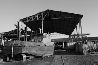 An old boat is positioned on a set of tracks under a large, metal-framed roof structure in a dockyard. The area appears industrial with a combination of open spaces and corrugated metal buildings. The ground is rough and scattered with debris, adding a sense of abandonment or disuse to the scene.