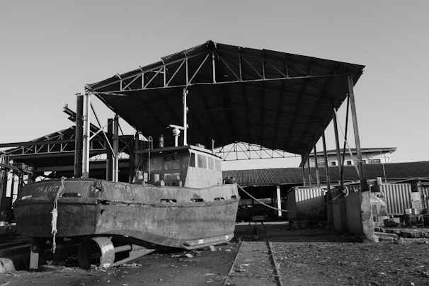 An old boat is positioned on a set of tracks under a large, metal-framed roof structure in a dockyard. The area appears industrial with a combination of open spaces and corrugated metal buildings. The ground is rough and scattered with debris, adding a sense of abandonment or disuse to the scene.