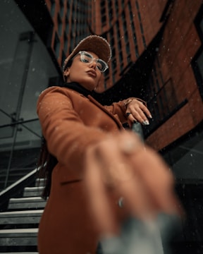 A person wearing a brown coat and cap stands confidently with a building in the background. The person is looking up slightly, wearing glasses and displaying an assertive expression.