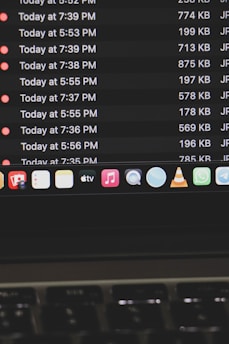 A computer screen displays a list of files with details such as timestamps, file sizes, and formats in a dark-themed interface. The bottom part of the screen shows various application icons on a MacOS dock, including familiar apps like VLC, WhatsApp, and Apple Music. A keyboard is partially visible at the bottom.
