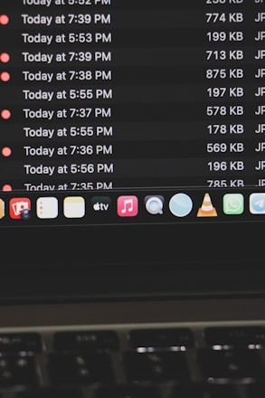 A computer screen displays a list of files with details such as timestamps, file sizes, and formats in a dark-themed interface. The bottom part of the screen shows various application icons on a MacOS dock, including familiar apps like VLC, WhatsApp, and Apple Music. A keyboard is partially visible at the bottom.