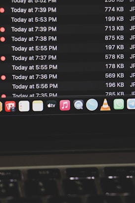 A computer screen displays a list of files with details such as timestamps, file sizes, and formats in a dark-themed interface. The bottom part of the screen shows various application icons on a MacOS dock, including familiar apps like VLC, WhatsApp, and Apple Music. A keyboard is partially visible at the bottom.