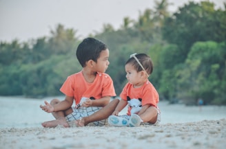 Two young children, possibly siblings, sit together on a beach. Both are wearing matching orange shirts with a print of a cute animal face. The child on the left is slightly older, while the one on the right wears a headband. They are sitting on the sandy shore with a backdrop of lush green trees and calm water.