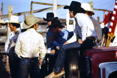 A group of men wearing cowboy hats and western-style clothing are gathered together outdoors. Some of them appear to be engaged in conversation, with one man sitting on the side of a vehicle. There are red, white, and black flags visible in the background, along with wooden fencing and a clear blue sky.