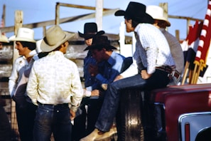 A group of men wearing cowboy hats and western-style clothing are gathered together outdoors. Some of them appear to be engaged in conversation, with one man sitting on the side of a vehicle. There are red, white, and black flags visible in the background, along with wooden fencing and a clear blue sky.