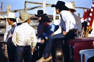 A group of men wearing cowboy hats and western-style clothing are gathered together outdoors. Some of them appear to be engaged in conversation, with one man sitting on the side of a vehicle. There are red, white, and black flags visible in the background, along with wooden fencing and a clear blue sky.