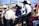 A group of men wearing cowboy hats and western-style clothing are gathered together outdoors. Some of them appear to be engaged in conversation, with one man sitting on the side of a vehicle. There are red, white, and black flags visible in the background, along with wooden fencing and a clear blue sky.