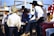 A group of men wearing cowboy hats and western-style clothing are gathered together outdoors. Some of them appear to be engaged in conversation, with one man sitting on the side of a vehicle. There are red, white, and black flags visible in the background, along with wooden fencing and a clear blue sky.