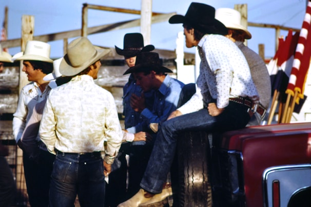 A group of men wearing cowboy hats and western-style clothing are gathered together outdoors. Some of them appear to be engaged in conversation, with one man sitting on the side of a vehicle. There are red, white, and black flags visible in the background, along with wooden fencing and a clear blue sky.