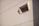 A single, small, rectangular window inset into a beige concrete or stucco wall with visible lines, casting a shadow to the side. The window has a protruding frame and seems to be made of glass with a grid pattern.