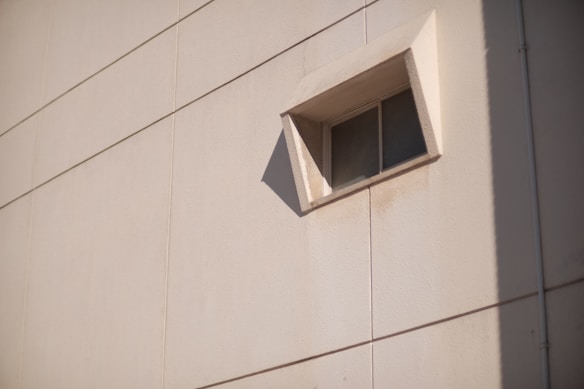 A single, small, rectangular window inset into a beige concrete or stucco wall with visible lines, casting a shadow to the side. The window has a protruding frame and seems to be made of glass with a grid pattern.