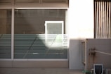 A glass window with horizontal frosted stripes and a light source casting a bright rectangle of light onto it. Surrounding the window is a white wall with some shadows. To the right, there's a concrete wall with a hose reel attached.
