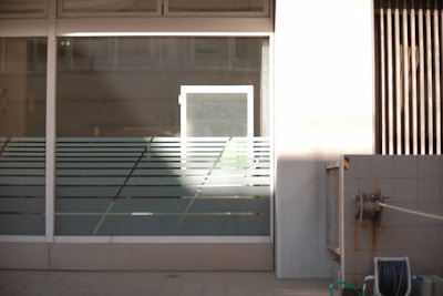 A glass window with horizontal frosted stripes and a light source casting a bright rectangle of light onto it. Surrounding the window is a white wall with some shadows. To the right, there's a concrete wall with a hose reel attached.
