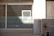 A glass window with horizontal frosted stripes and a light source casting a bright rectangle of light onto it. Surrounding the window is a white wall with some shadows. To the right, there's a concrete wall with a hose reel attached.
