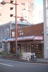 A small brick and wood storefront with a red awning displaying white text, situated along a quiet street. A bicycle is parked in front of the shop, and some potted plants line the entrance. A tall street lamp is positioned in the foreground, with industrial buildings visible in the background.