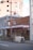 A small brick and wood storefront with a red awning displaying white text, situated along a quiet street. A bicycle is parked in front of the shop, and some potted plants line the entrance. A tall street lamp is positioned in the foreground, with industrial buildings visible in the background.