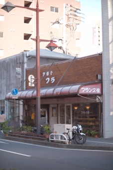 A small brick and wood storefront with a red awning displaying white text, situated along a quiet street. A bicycle is parked in front of the shop, and some potted plants line the entrance. A tall street lamp is positioned in the foreground, with industrial buildings visible in the background.