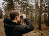 A person is standing in a forested area, holding binoculars up to their eyes. The background consists of tall trees with brown trunks and sparse green foliage. The person is wearing a black jacket.